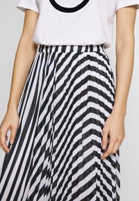 Kvinna i en vit t-shirt instoppad i en högmidjad plisserad kjol med svart- och vit diagonala ränder.