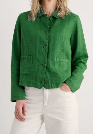 Frau trägt eine grüne, geknöpfte Jacke mit zwei vorderen Taschen und weiße Hose, eine Hand in der Tasche, vor schlichtem Hintergrund.