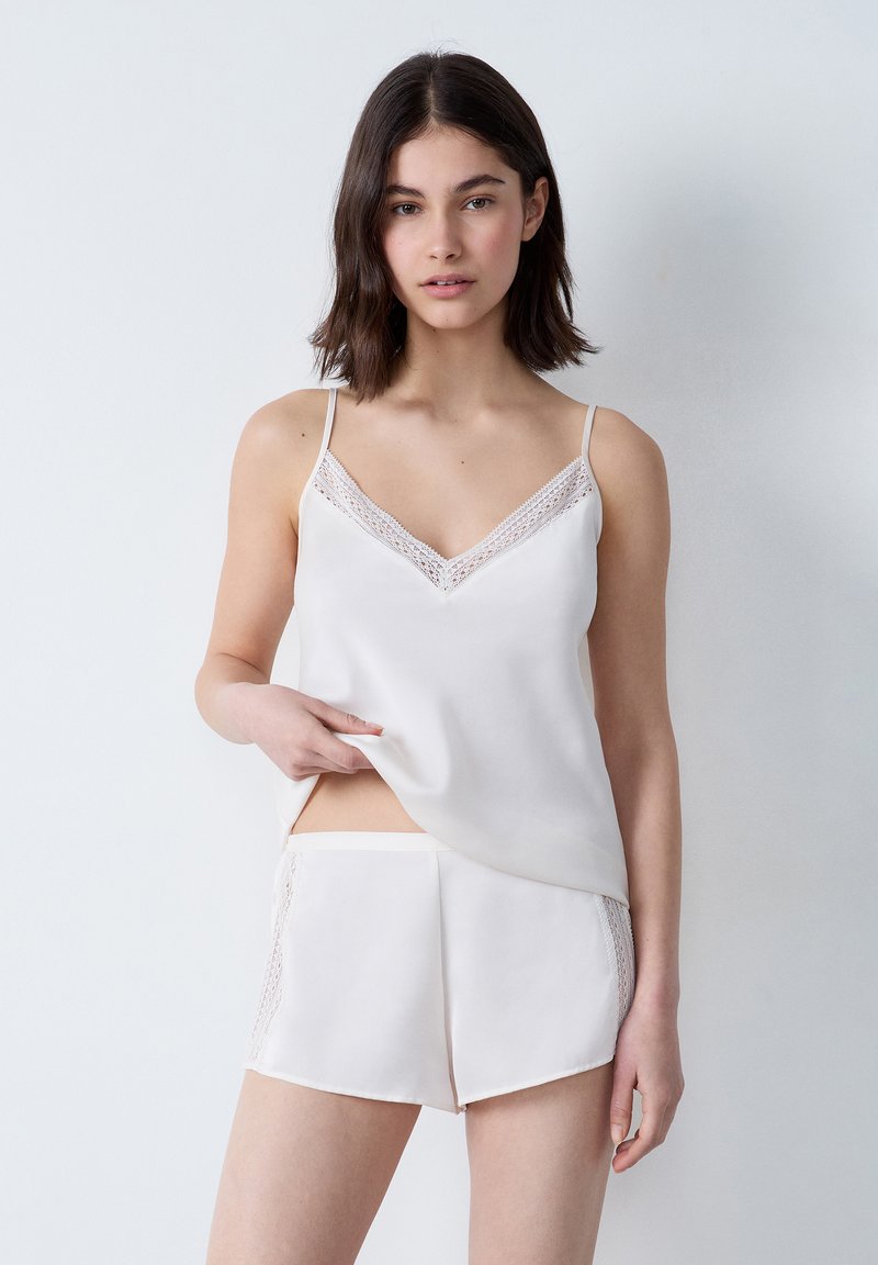 Zijdeachtige witte camisol met kant langs de V-hals, gecombineerd met bijpassende shorts met kanten accenten aan de zijkanten.