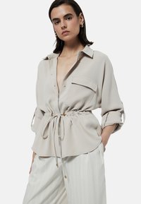Lichtbeige blouse met een kraag, trekkoord in de taille en lange mouwen opgerold. Heeft een zak aan de linkerkant en een gladde textuur.