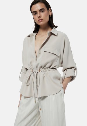 Blouse beige clair avec un col, taille à cordon de serrage et manches longues retroussées. Présente une poche sur le côté gauche et une texture lisse.