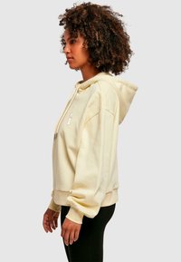 Sudadera con capucha amarilla, con bolsillo canguro, capucha con cordón y puños acanalados. Presenta un diseño bordado en el pecho.