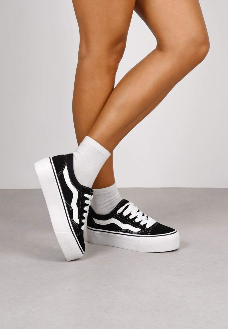 Zapatillas de lona negras con detalles blancos y suelas de goma, que cuentan con un diseño de cordones y un perfil bajo. Se ven calcetines blancos lisos.