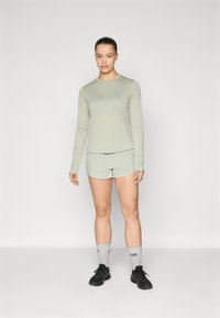 Un haut à manches longues vert clair et un short assorti avec une texture lisse. La tenue présente un logo discret et est associée à des chaussures noires et des chaussettes grises.