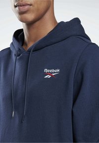 Hoodie bleu marine en mélange de coton doux, doté d'une capuche avec cordon de serrage et d'un logo Reebok brodé en blanc et rouge sur la poitrine.