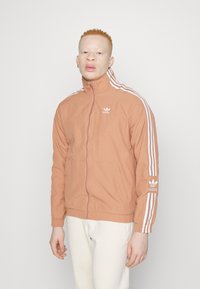 Persiku krāsas jaku ar rāvējslēdzēju un baltām Adidas svītrām un logo. Tā ir ar augstu apkakli un sānu kabatām. Valkāta virs gaišiem biksēm.