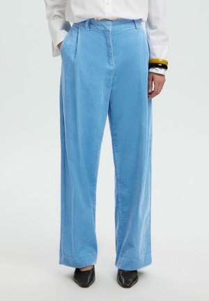 Personne portant un pantalon large en velours côtelé bleu clair, une chemise blanche, des chaussures noires pointues et des bracelets épais jaunes et noirs à un poignet.