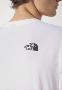 Camiseta de algodón blanca con un pequeño logo negro de diseño arqueado en la parte superior de la espalda, que muestra una textura suave y un ajuste casual.