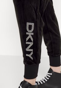 Underbenet bär svarta velourbyxor med "DKNY" i rhinestones och svarta läderhög-toppsneakers med vita snörningar.