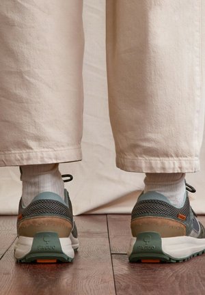 Pieds chaussés de baskets Faguo gris et marron, avec des chaussettes blanches et un pantalon beige, debout sur un sol en bois.
