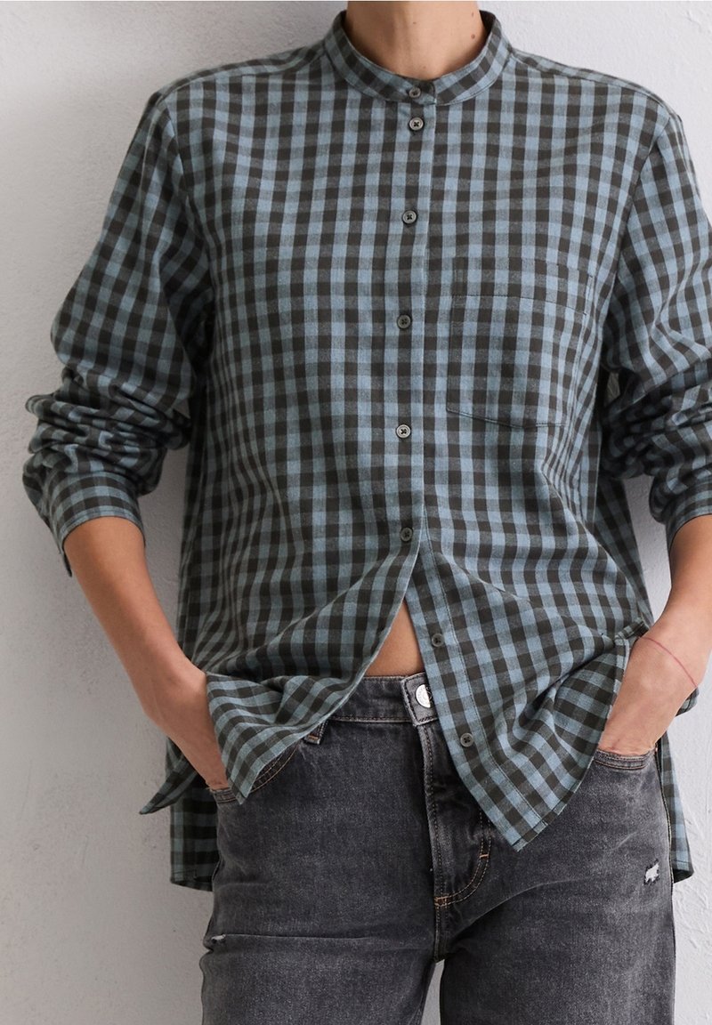 Personne portant une chemise à carreaux bleue et noire boutonnée, manches retroussées, mains dans les poches d'un jean noir délavé.