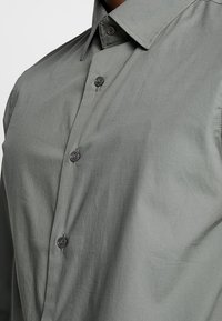 Koszula z guzikami w jasnoszarym materiale, z kołnierzem rozłożystym, sześcioma guzikami i gładkim, teksturowanym wykończeniem.