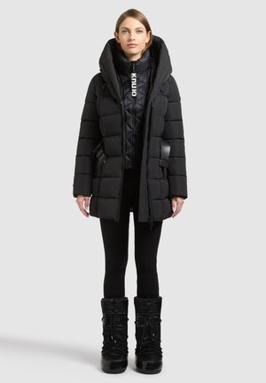 Mujer con un abrigo largo de invierno negro acolchado con capucha grande, pantalones negros y botas negras de invierno con cordones, de pie frente a un fondo liso.