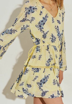 Robe florale courte jaune avec des fleurs bleues, décolleté en V, manches longues évasées, taille cintrée avec un lien jaune, et détails en dentelle au bas.