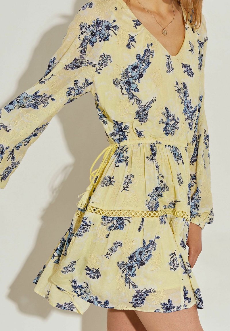 Vestido corto amarillo con flores azules, escote en v, mangas largas acampanadas, cintura ajustada con un lazo amarillo y detalles de encaje en el dobladillo.
