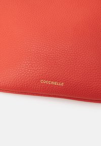 Zbliżenie teksturowanej powierzchni czerwonej skóry z małą, złotą wytłoczoną nazwą marki "Coccinelle" na dole pośrodku.