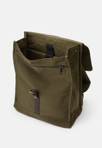 Sac à dos en tissu vert olive avec un grand compartiment principal, une poche zippée à l'avant et un accent de sangle marron. Dispose de bretelles réglables.