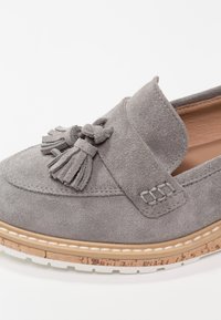 Chaussure en suède gris avec un accent de pampille, un bout arrondi et une semelle intérieure rembourrée. Elle présente une base en liège texturée et une semelle extérieure en caoutchouc blanc.