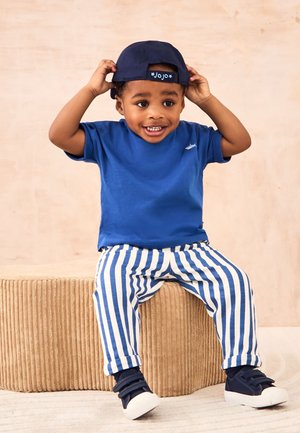 Småbarn iført en blå skjorte, stribede bukser, sneakers og en bagudvendt marineblå kasket sidder på en struktureret beige puf og smiler.