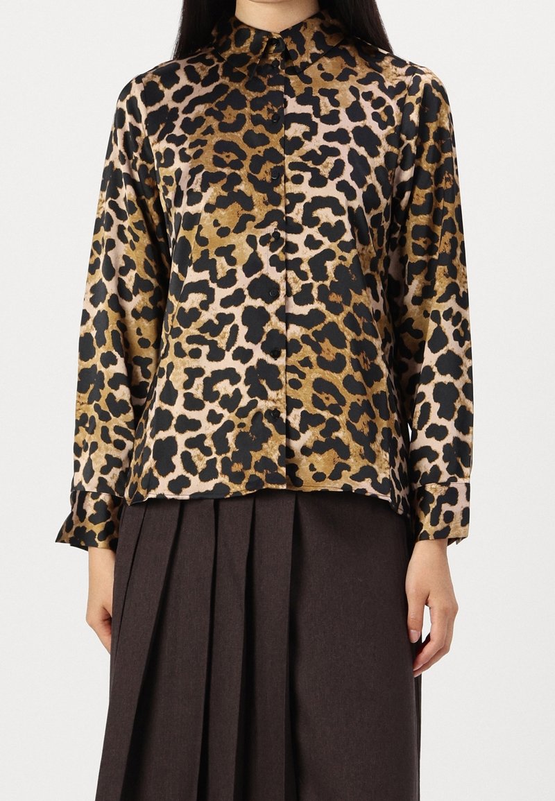 Personne portant une chemise à manches longues boutonnée avec imprimé léopard et une jupe plissée foncée sur un fond uni.