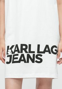 Balta kokvilnas T-kreklu kleita ar izteiksmīgu melnu tekstu "KARL LAGERFELD JEANS" un nelielu grafisku detaļu, ar brīvu piegriezumu un līdz ceļiem garumu.