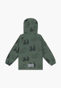 Grüne Kinderjacke mit Kapuze, langen Ärmeln und einem schwarzen Truck-Muster. Auffällige Details sind ein strukturierter Patch mit Löchern.