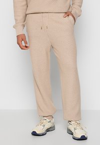Beige knitted pants with a textured pattern, featuring an elastic waistband and drawstring. Paired with cream athletic shoes with blue accents.