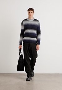 Strickpullover mit einem Farbverlauf von Marineblau zu Hellgrau gestreift, kombiniert mit schwarzen Hosen und einer schwarzen Tote-Tasche. Glatte Textur und gerippter Kragen.