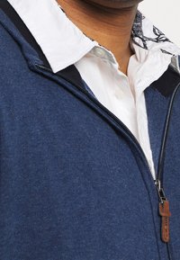 Námořnicky modrá mikina na zip vyrobená z texturované tkaniny, s bílou košilí pod ní. Obsahuje kožený táhlo na zip a kontrastní černé detaily.