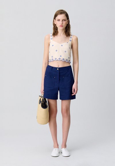 Débardeur crème court avec des motifs en losanges bleus, associé à un short bleu marine avec des poches avant, et tenant un sac rond en paille.