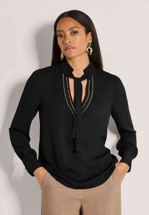 Vrouw draagt een zwarte blouse met lange mouwen met een met studs versierde halslijn en strikdetail, gecombineerd met beige broek en grote gouden oorbellen.