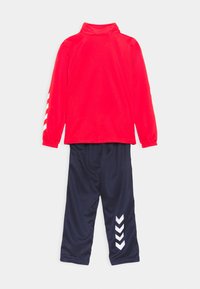 Röd långärmad topp med hög krage och vita accenter; kombinerad med marinblå byxor med vita chevronmönster på sidorna.