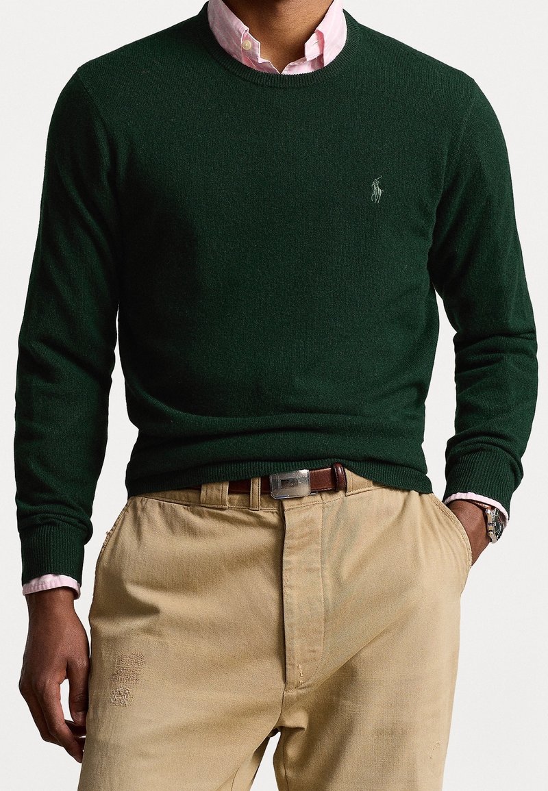 Man draagt een donkergroene trui met ronde hals over een roze overhemd, beige broek met een bruine riem en een horloge, handen in de zakken tegen een effen achtergrond.