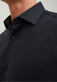Camisa negra de botones con collar extendido, textura suave y un brillo sutil, con un solo botón visible en la tapeta.