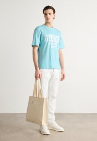 Lyseblå bomulls t-skjorte med "HUGO CREATIVE DEPT." i hvitt, kombinert med hvite jeans og en beige tote-sekk med skulderremmer.