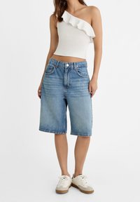 Witte top met één schouder en ruches, gecombineerd met lichtblauwe denim shortjes tot halverwege het bovenbeen en witte sneakers met bruine accenten.
