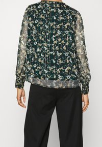 Femme portant un chemisier à manches longues en tissu transparent avec un imprimé floral abstrait en vert foncé, beige et blanc, associé à un pantalon noir ample, vue de dos.