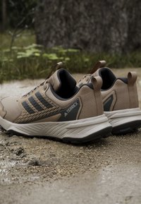 Tan waterproof athletic shoes with black accents and textured fabric, featuring a rubber sole, looped heel tab, and three stripes design.