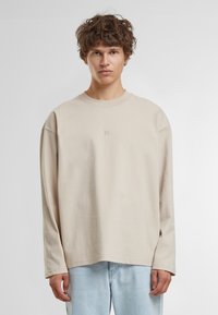 Beige Langarm-T-Shirt mit Rundhalsausschnitt und überschnittenen Schultern, versehen mit einem kleinen gestickten Logo auf der Brust. Weiches Baumwollmaterial.