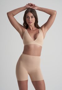 Kvinna står med armarna lyfta och händerna knutna ovanför huvudet, iförd en beige sömlös bh och höga formande shorts mot en enfärgad bakgrund.