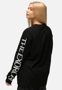 Svart långärmad t-shirt med vit text "THE EXORCIST" längs med vänster ärm. Tillverkad av mjukt tyg med en avslappnad passform.