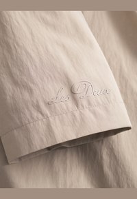 Materialul de culoare bej deschis are o suprafață texturată, cu un logo brodat "Les Deux" pe margine. Designul este simplu și minimal.