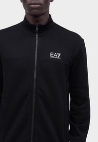 Homme portant une veste noire zippée avec un col montant et un logo EA7 Emporio Armani blanc sur la poitrine gauche.