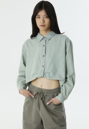 Camicetta verde chiaro corta con bottoni frontali e colletto, abbinata a pantaloni beige larghi con vita regolabile. Tessuto con trama.