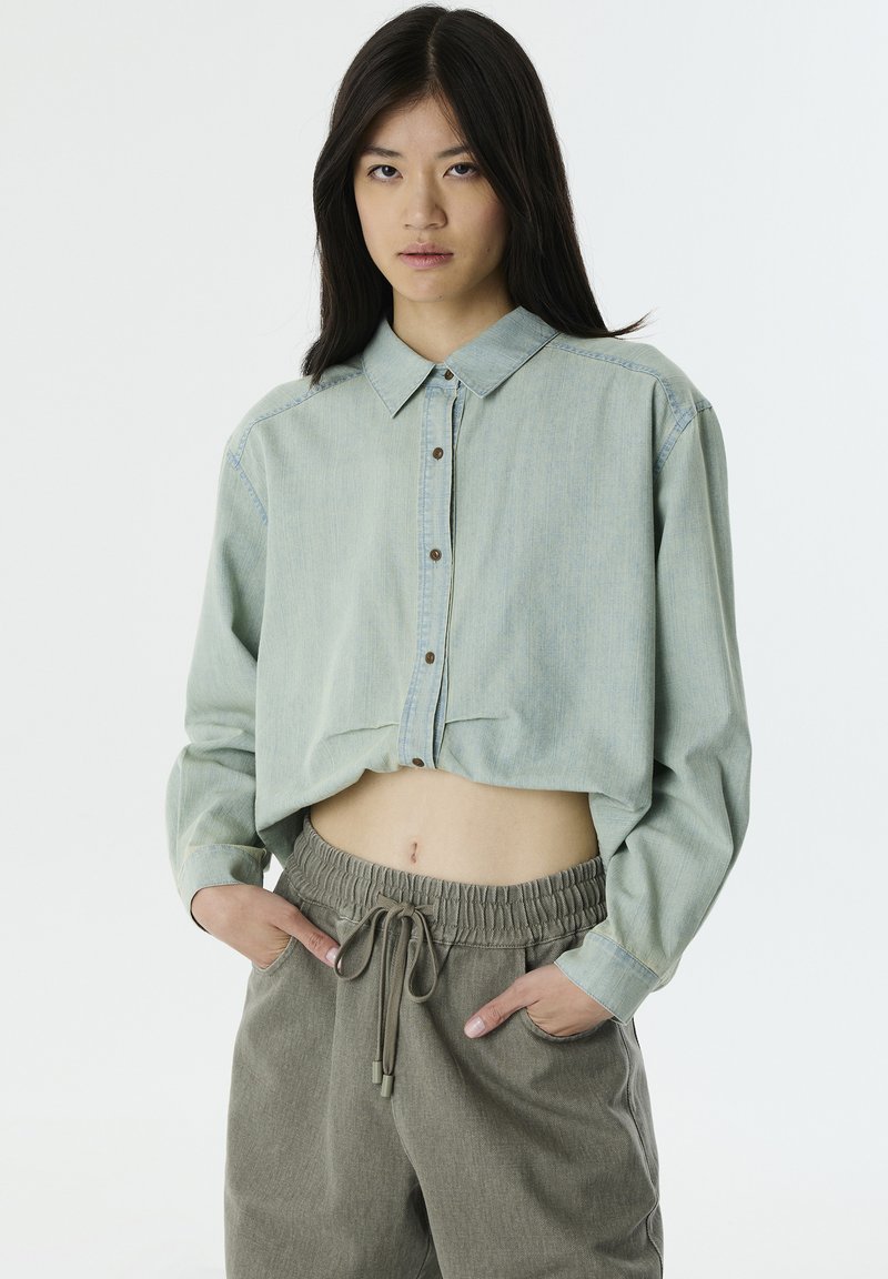 Camicetta verde chiaro corta con bottoni frontali e colletto, abbinata a pantaloni beige larghi con vita regolabile. Tessuto con trama.