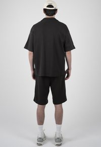 Homme portant une chemise noire à manches courtes et un short, casquette blanche et baskets blanches, debout de dos devant un fond gris uni.
