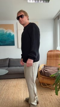 Black knit sweater, beige trousers, and gray shoes; rectangular sunglasses; indoor setting with a gray couch and rattan chair visible.