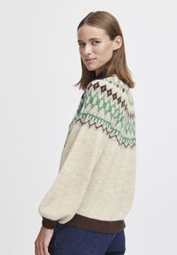 Knit sweater in cream with green and brown geometric patterns. Features a round neckline and ribbed cuffs. Soft texture, loose fit.