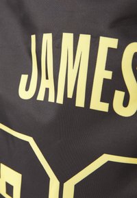 Superfície de tecido preto com grandes letras amarelas que dizem "JAMES" com um padrão geométrico contornado em amarelo. Acabamento texturizado.