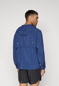 Μπλε-ναυτικό μπουφάν με κουκούλα από ελαφρύ ύφασμα, με πίσω άνοιγμα, ρυθμιζόμενες μανσέτες και ανακλαστικές λεπτομέρειες. Φοριέται με μαύρη βερμούδα.